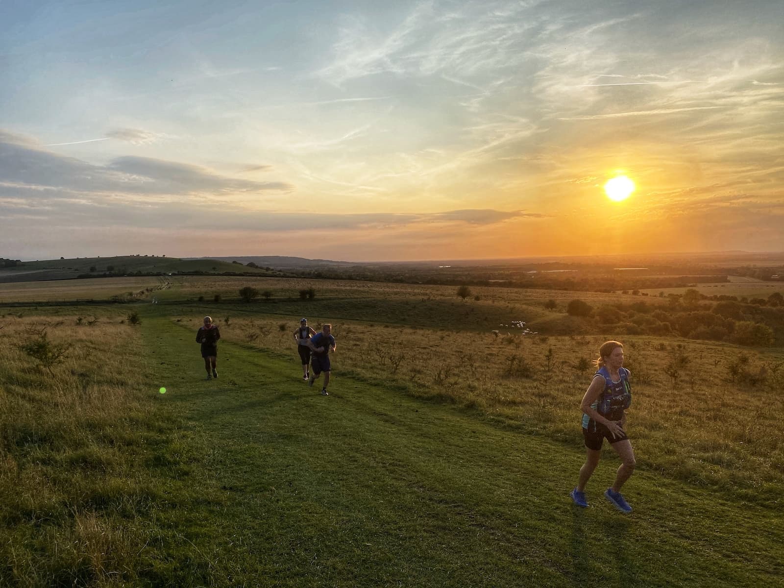 Four people are trail running on a grassy path through a wide, open field during sunset. The sky is vibrant with orange and yellow hues as the sun sets on the horizon. Rolling hills and scattered trees can be seen in the distance.