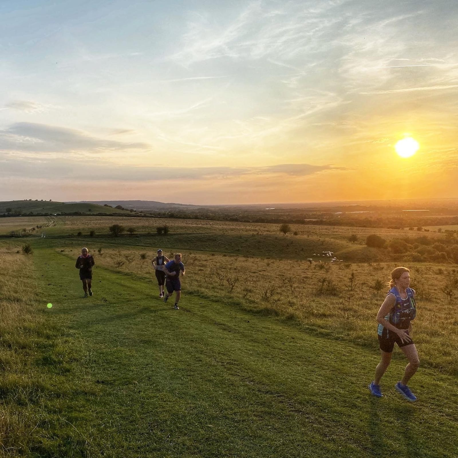 People are running on a grassy path through rolling hills during sunset. The landscape is bathed in the warm golden light of the setting sun, creating a serene and peaceful atmosphere. The sky is mostly clear with a few scattered clouds.