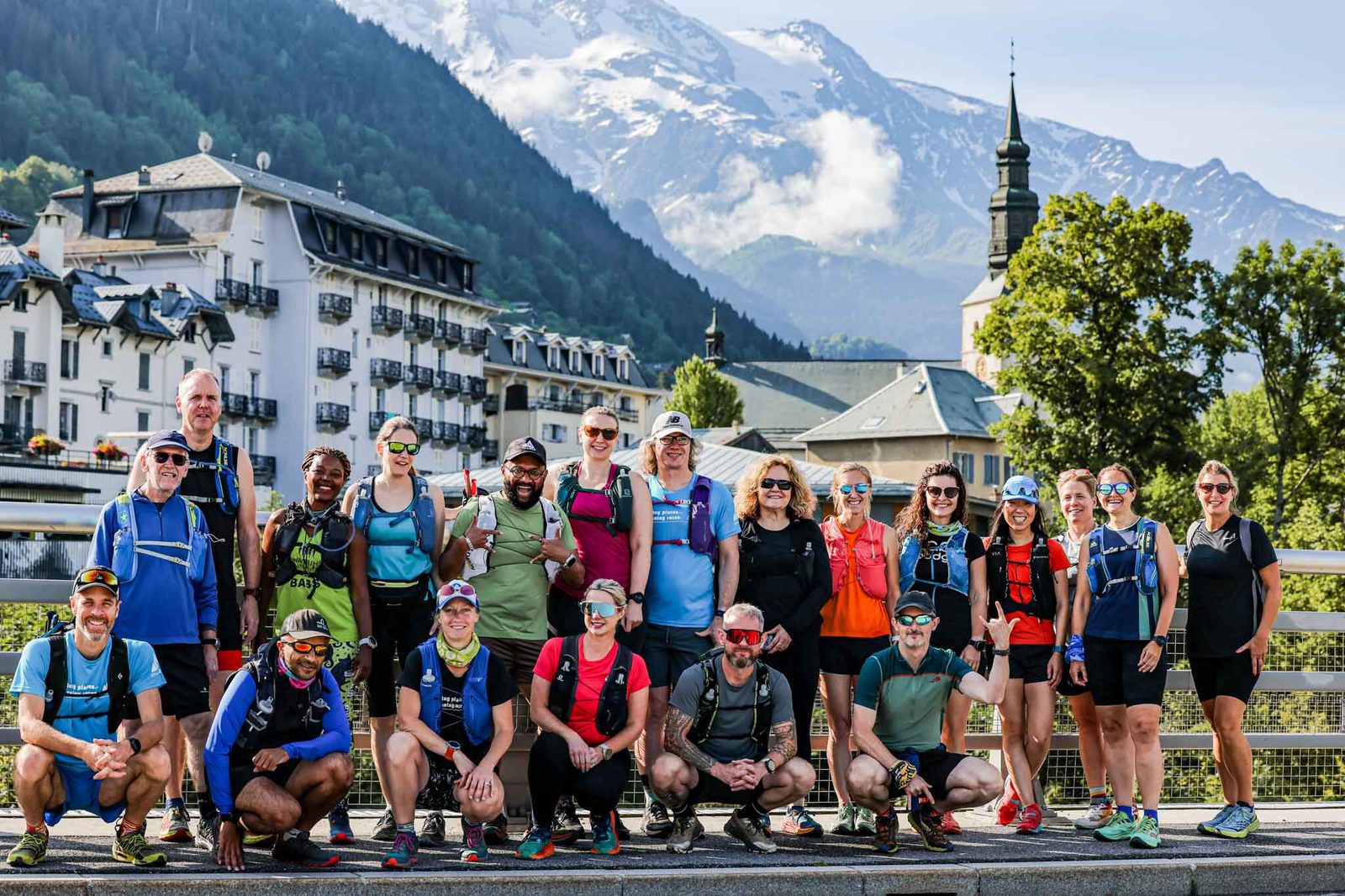 A diverse group of people in athletic gear pose for a photo on a bridge with a scenic mountain backdrop. They are smiling, appearing ready for an outdoor adventure. The scene includes a picturesque village with charming buildings and a church tower.