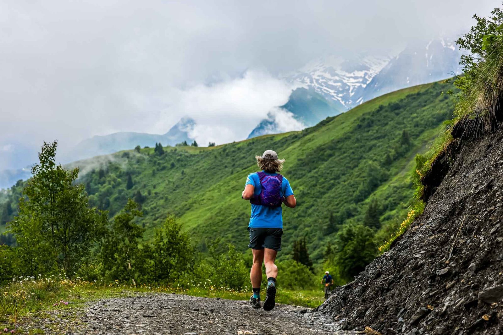 A person with gray hair, wearing a blue shirt and purple backpack, runs along a dirt trail in a lush, green mountainous area. Mist and clouds partially cover the distant peaks, creating a serene and refreshing scene.