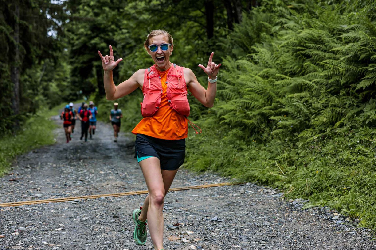 A woman in an orange tank top and sunglasses runs on a gravel trail through a forest, showing the "I love you" sign with both hands. A group of runners is visible in the background, and lush greenery surrounds the path.
