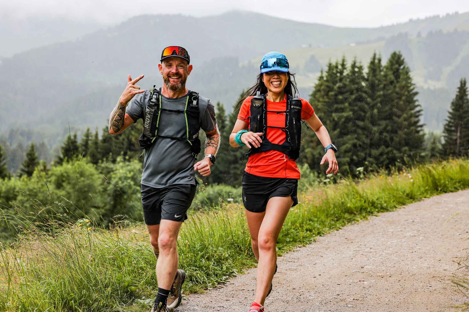 Two people are joyfully running on a dirt trail through a scenic, mountainous area surrounded by lush greenery and pine trees. The person on the left is smiling and making a hand gesture, while the person on the right, wearing a blue cap, is also smiling.