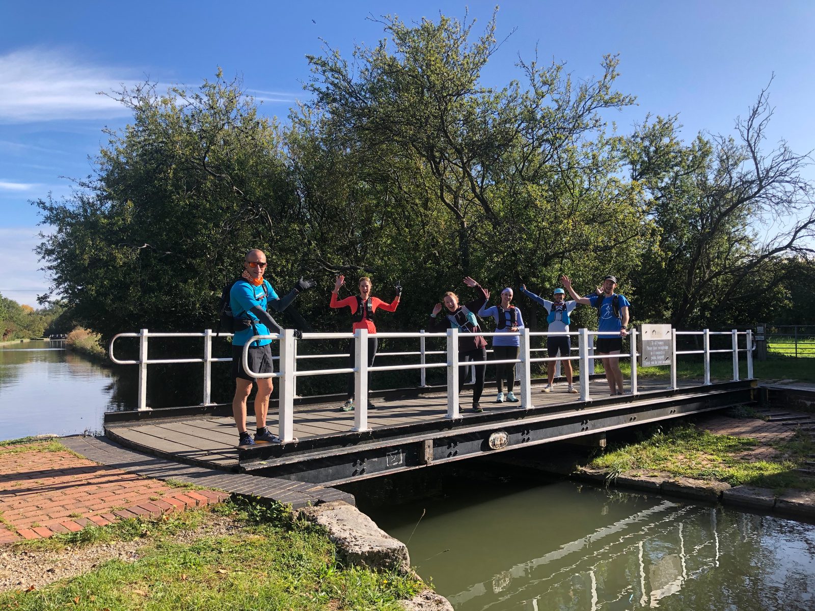 A group of six people stands and waves on a small bridge over a canal. They are dressed in casual outdoor attire, and the setting is a lush, green area with trees and clear skies. The atmosphere appears cheerful and relaxed.