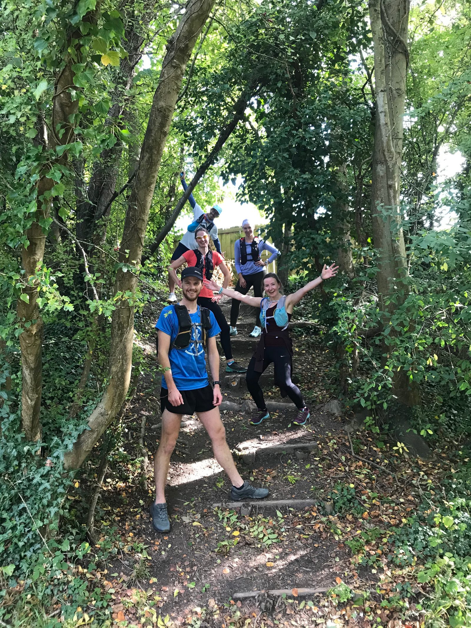 A group of five people dressed in athletic wear are standing on a leafy forest path, smiling, and striking playful poses. They appear to be on a hike or trail run, surrounded by lush green foliage and trees. The atmosphere looks cheerful and vibrant.