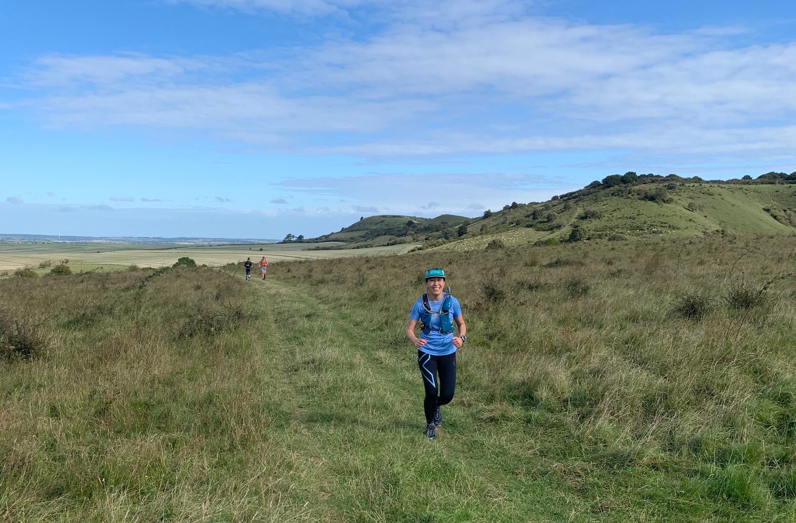 A person wearing a blue shirt and backpack jogs along a grassy trail through a hilly landscape under a blue sky with scattered clouds. In the distance, two other runners can be seen on the path. The scenery features rolling green hills and open fields.