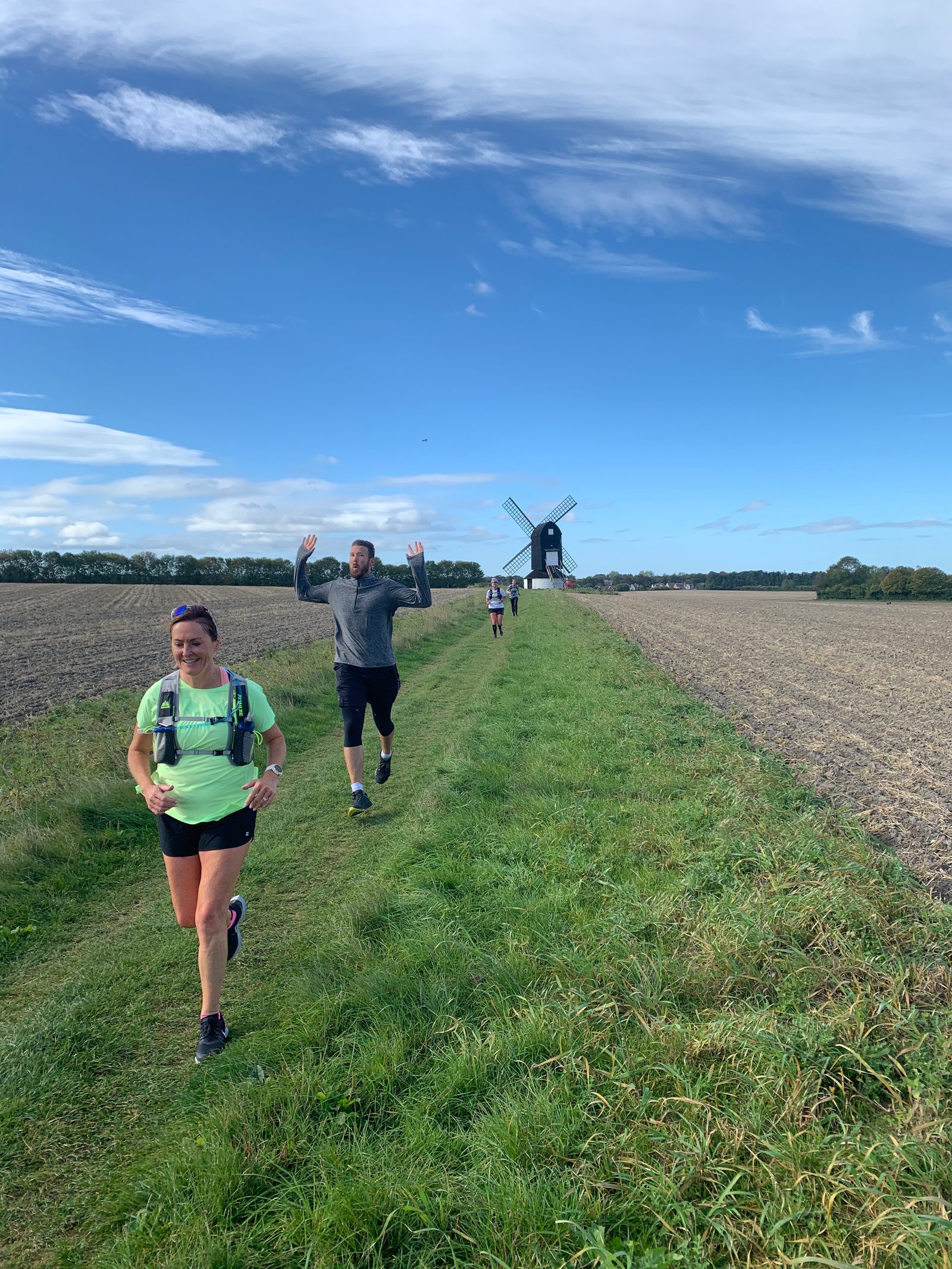 A woman and a man are running on a grassy path between two fields under a blue sky. The woman, in front, wears a light green shirt and a vest, while the man behind raises his hands cheerfully. A windmill is visible in the distance.