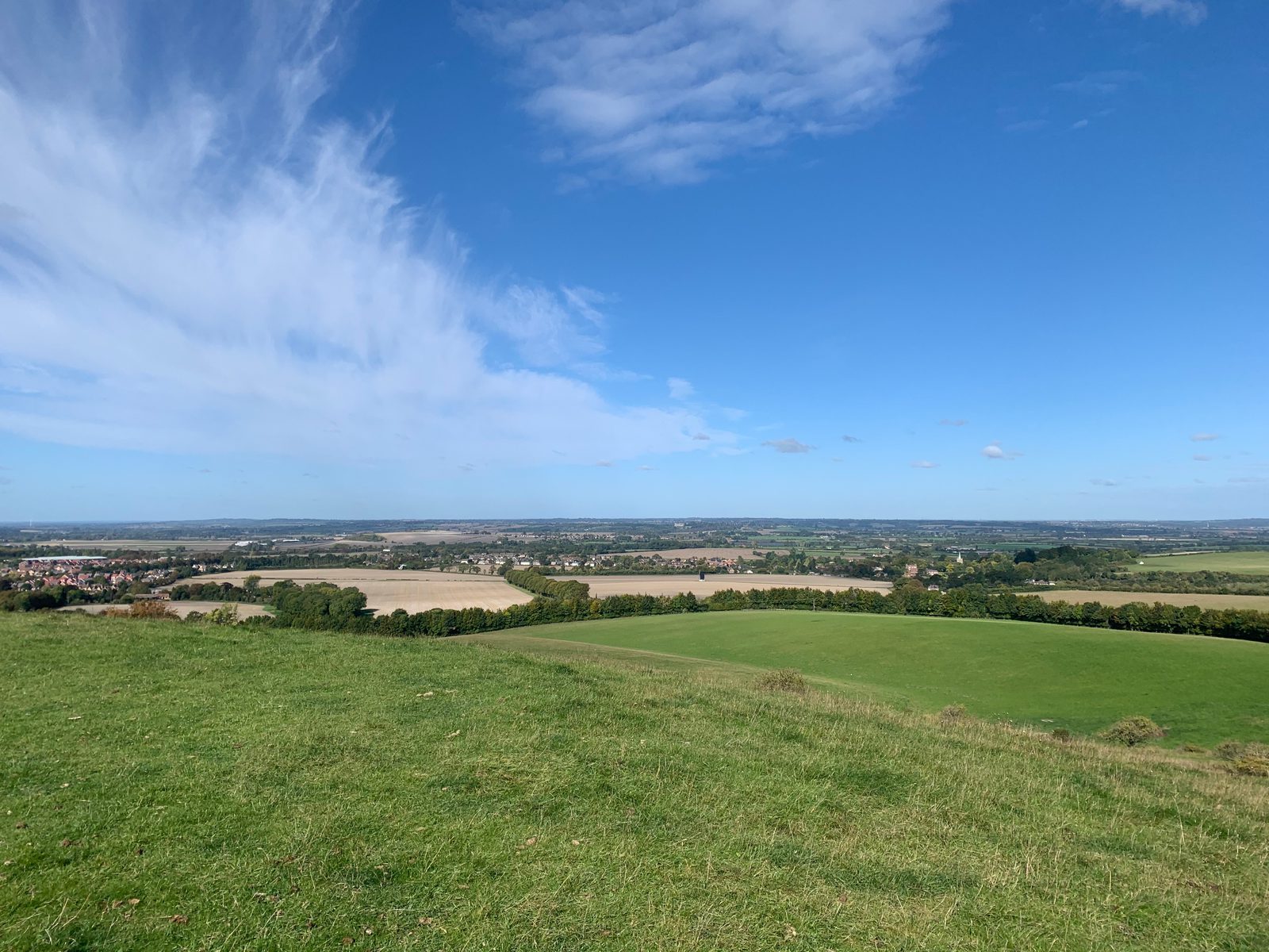 A scenic view from a grassy hilltop overlooking a vast expanse of fields and farmland under a blue sky with scattered clouds. The horizon shows a mix of greenery and small patches of settlement in the distance.