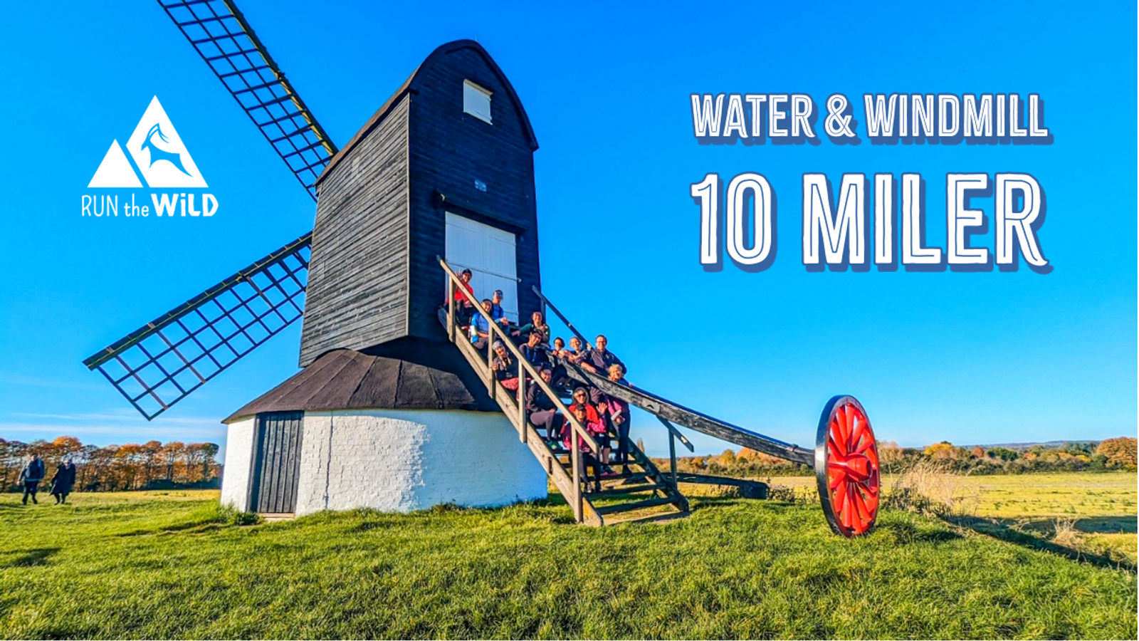 A group of people sits on a large wheel in front of a black windmill with a white base. The image includes text: "RUN the WILD," and "WATER & WINDMILL 10 MILER," on the right side against a clear blue sky.