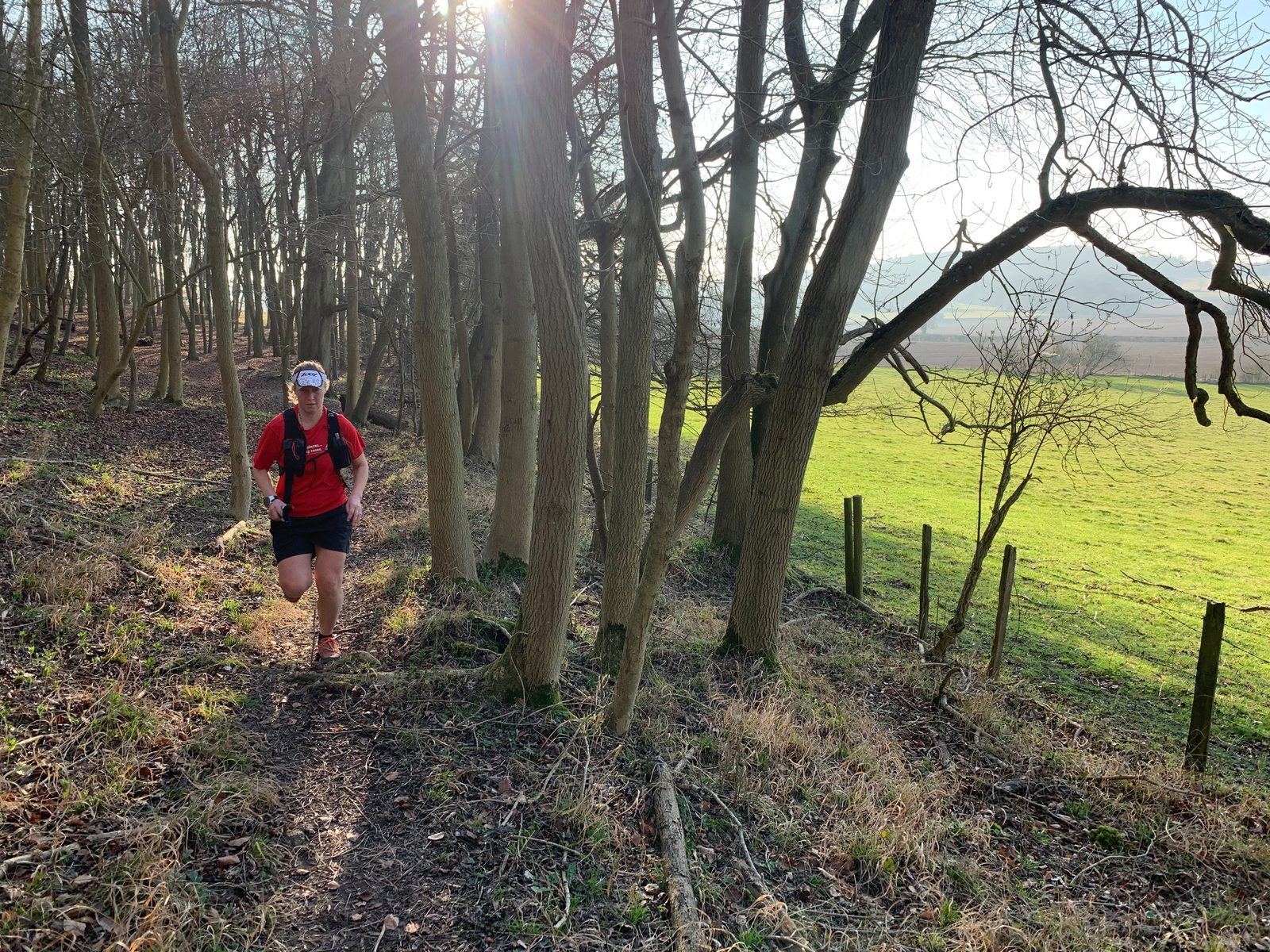 A person wearing a red shirt, black shorts, and a hat runs along a narrow path through a wooded area. Sunlight filters through the bare trees, and grassy fields are visible in the background. The day is bright and clear.
