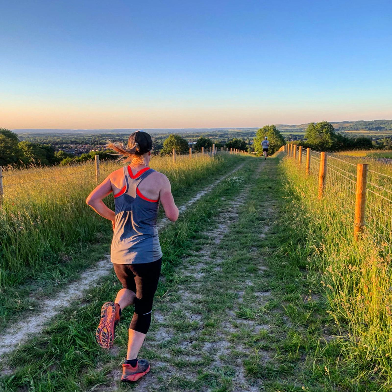 A person wearing a gray tank top, black leggings, and pink running shoes is running on a dirt path through a grassy field during sunset. The path is flanked by tall grass and fences, leading towards the horizon with trees and a clear sky in the background.