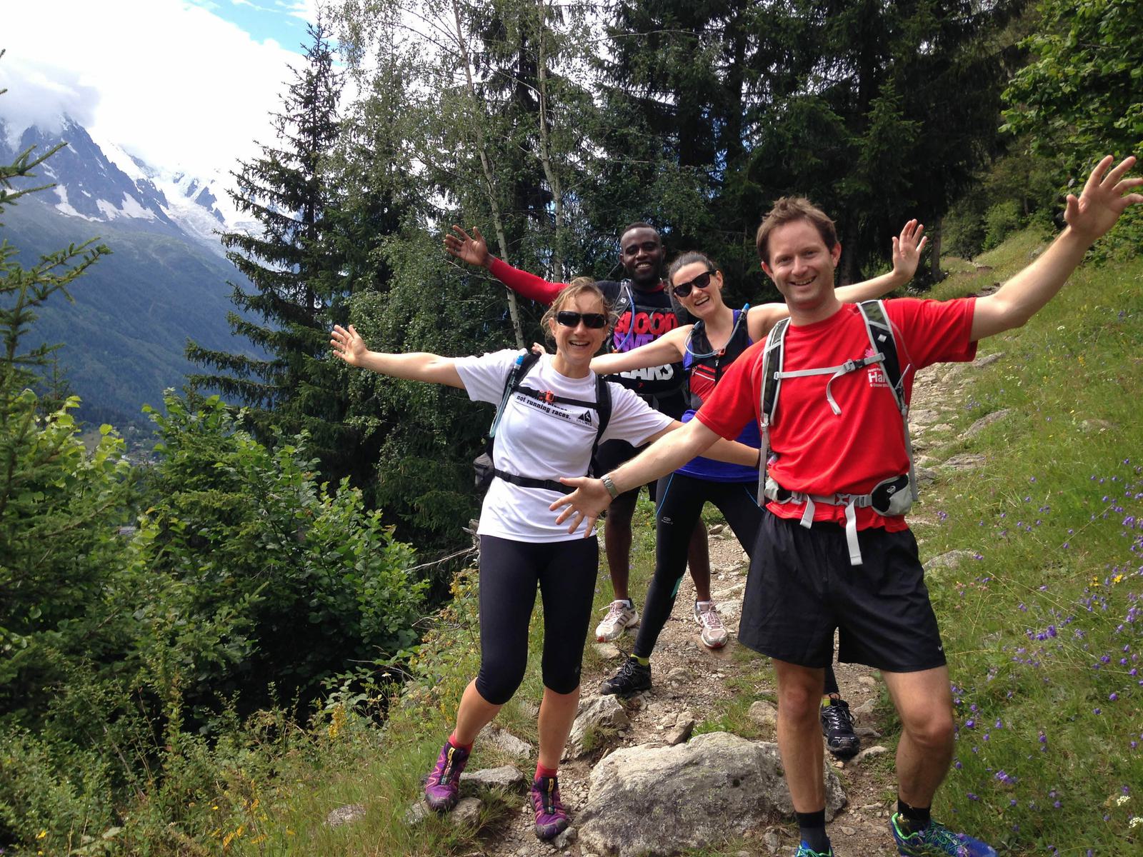Four people are outdoors on a hiking trail, posing energetically with arms spread out. They are dressed in athletic clothing with backpacks, surrounded by green trees and mountains in the background, under a partly cloudy sky.