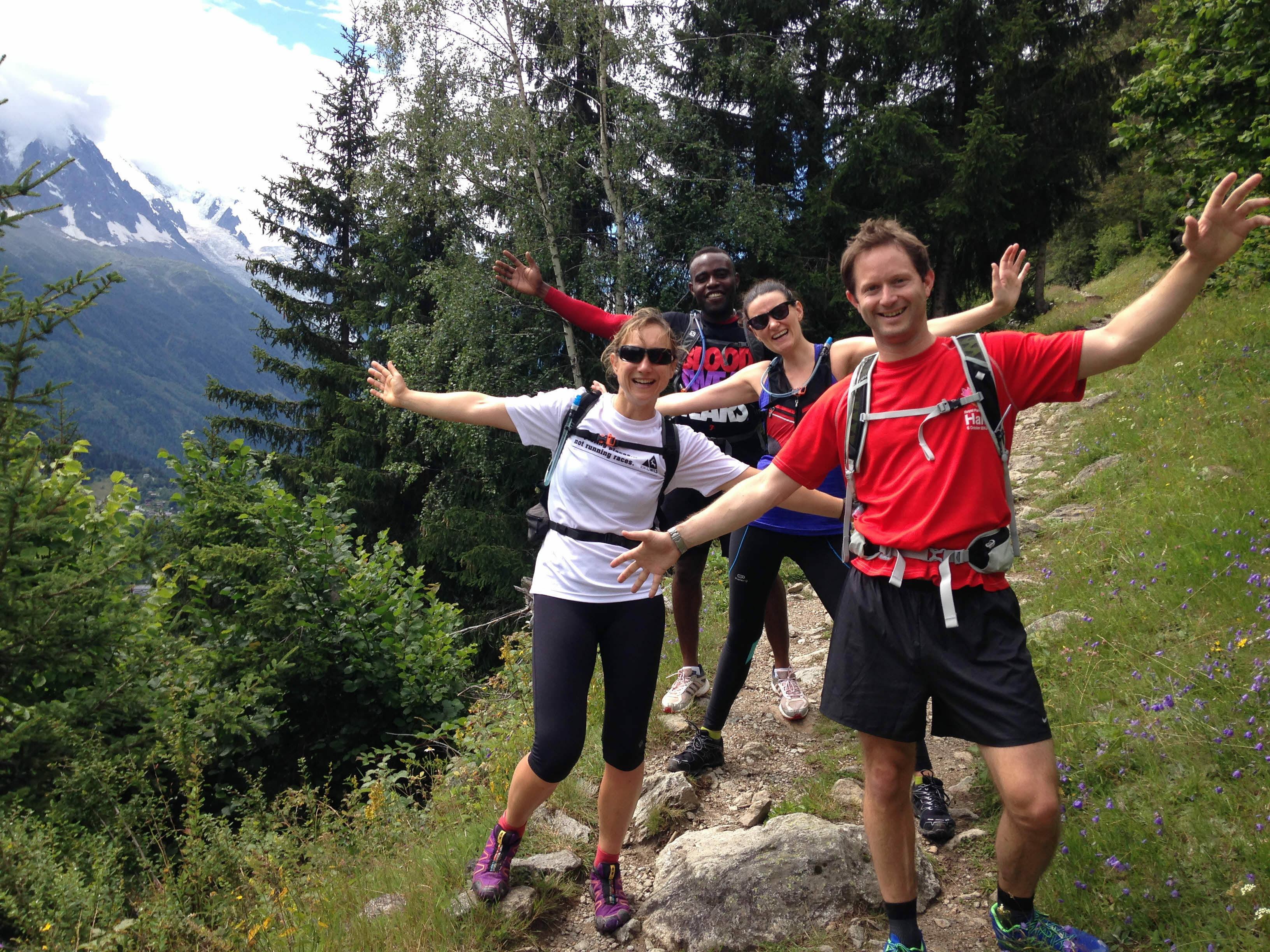 Four people are outdoors on a hiking trail, posing energetically with arms spread out. They are dressed in athletic clothing with backpacks, surrounded by green trees and mountains in the background, under a partly cloudy sky.