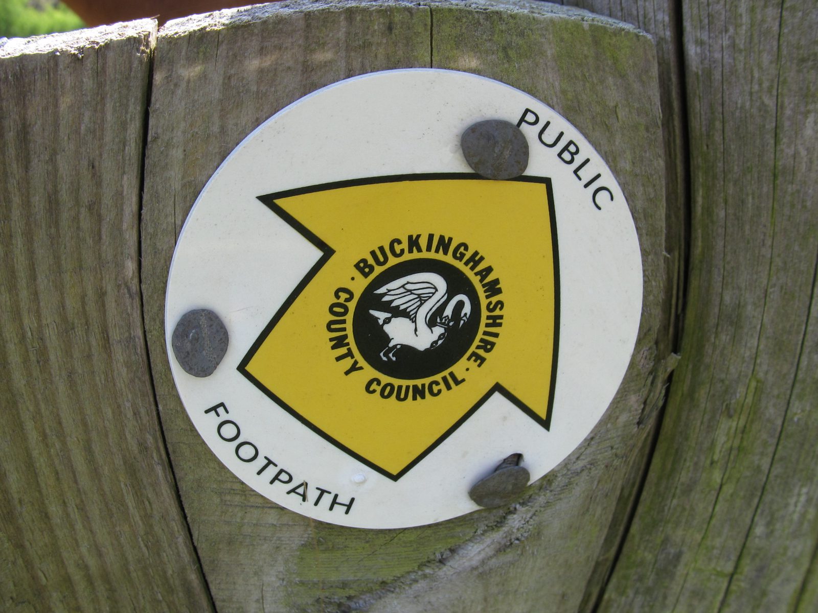 A yellow arrow sign mounted on a wooden surface indicates a public footpath in Buckinghamshire. The sign features the Buckinghamshire County Council emblem, which includes an image of a swan. The words "PUBLIC" and "FOOTPATH" are also visible.