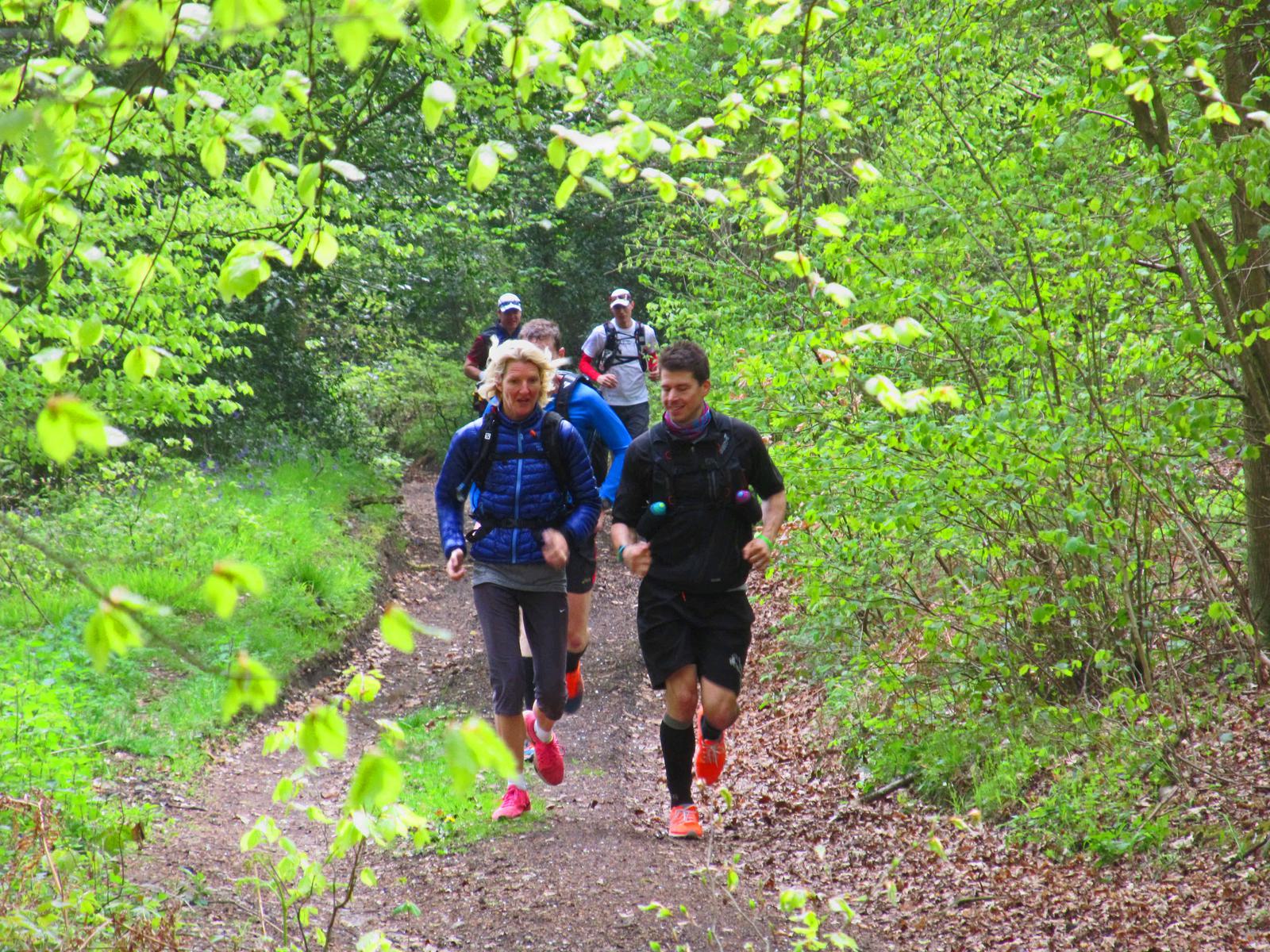 A group of runners jogs along a wooded trail, surrounded by lush green foliage. The leading pair, a man and a woman, both wear athletic outfits and appear focused. More runners follow behind them, partially visible through the trees. The scene is vibrant and energetic.