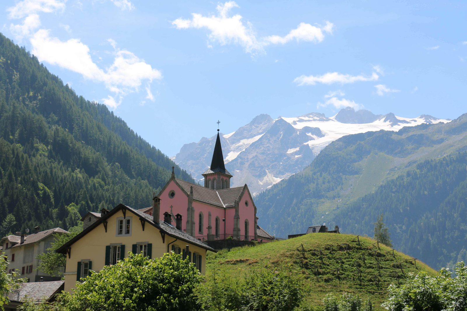 A quaint village nestled in the Alps, featuring a charming pink church with a black steeple as the centerpiece. Snow-capped mountains rise in the background under a partly cloudy sky, and green hills surround the village houses and trees.
