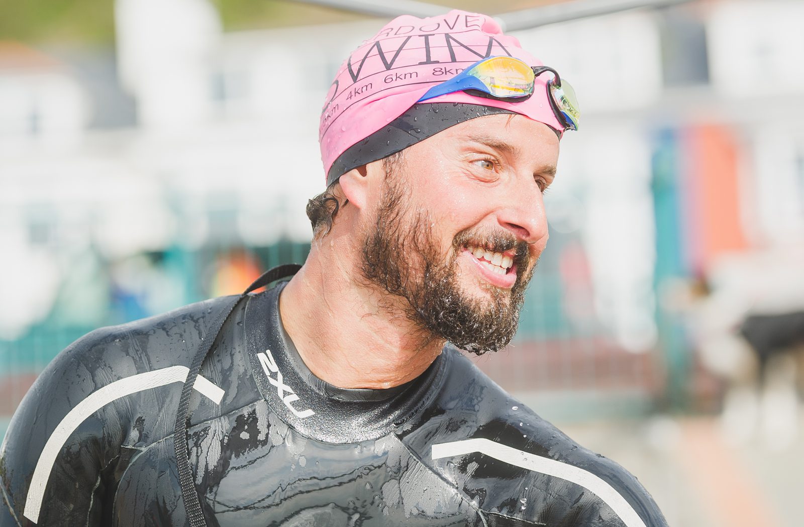 A man in a black wetsuit and a pink swim cap with goggles on top of the cap smiles cheerfully. He has a beard and appears to be wet, suggesting he just finished swimming. Buildings with colorful accents are blurred in the background.