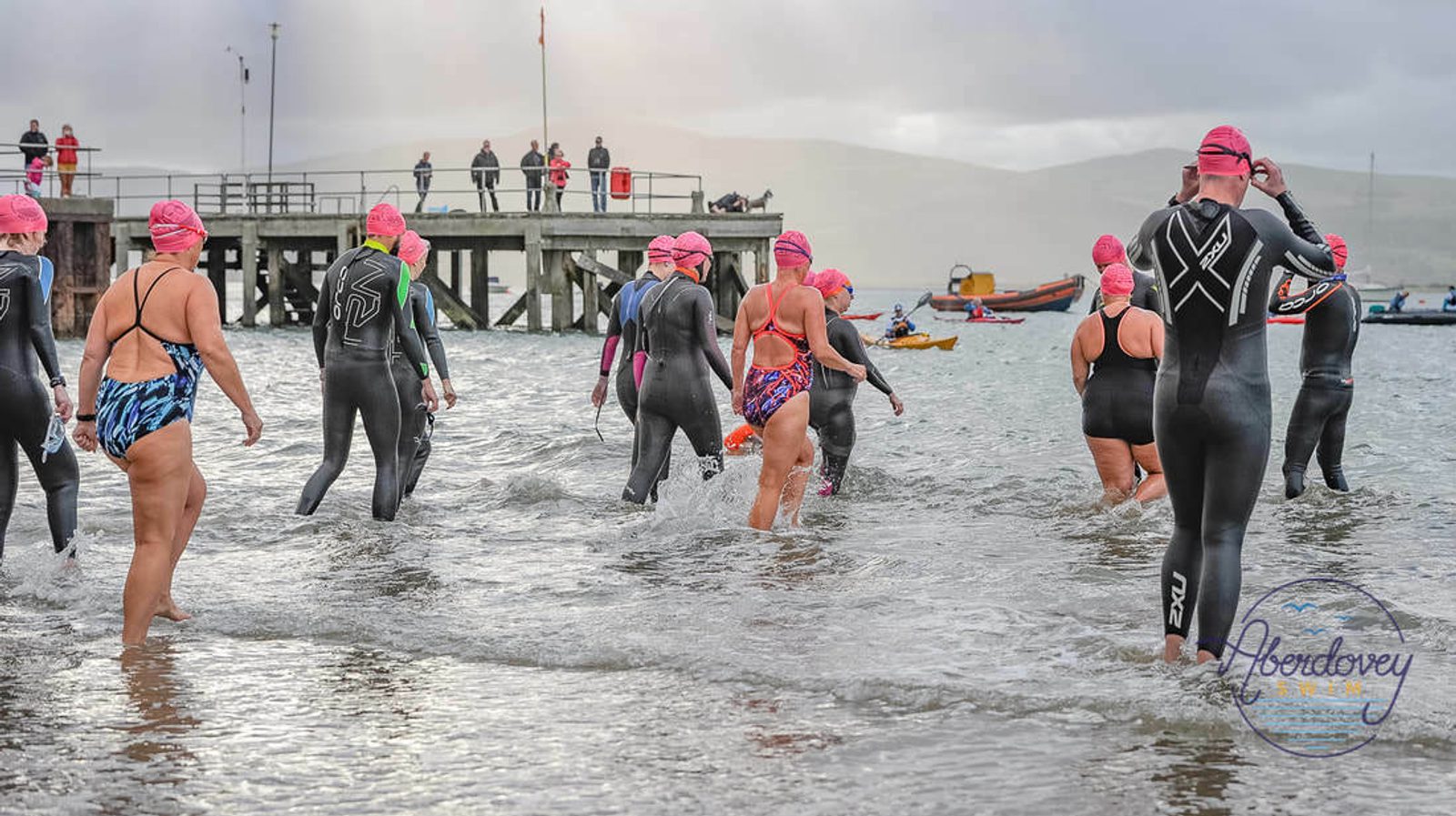 Swimmers wearing wetsuits and pink swim caps enter the water from the shore, preparing for a swim event. A pier with spectators and boats are visible in the background. The atmosphere appears overcast.