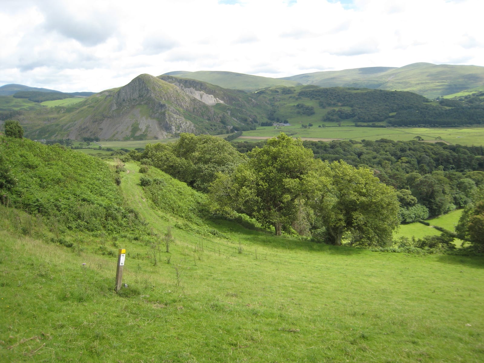 A scenic view of a lush green landscape with rolling hills and mountains in the background. The foreground features grassy meadows and a marked trail, with scattered trees and dense forest in the distance under a partly cloudy sky.