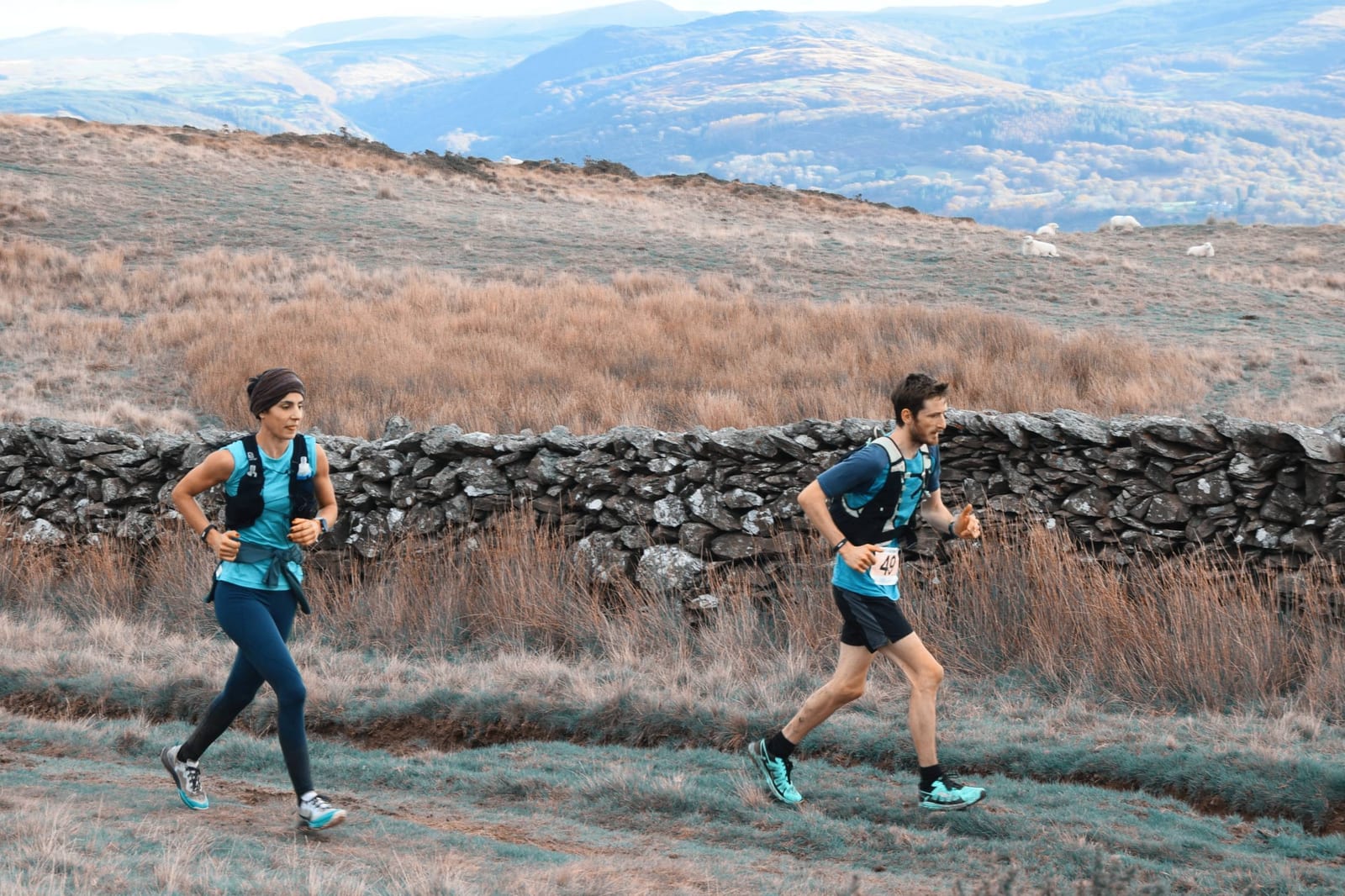 Two runners, wearing blue athletic gear and running vests, jog along a trail in a scenic mountainous area with dry grass, a stone wall, and rolling hills in the background. The sky is clear, and both runners appear focused and determined.