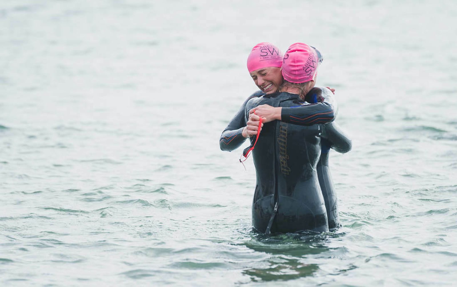 Two participants in black wetsuits and pink swim caps embrace in shallow water during a swimming event. Both appear joyful and relieved, sharing a celebratory hug in the middle of the water.