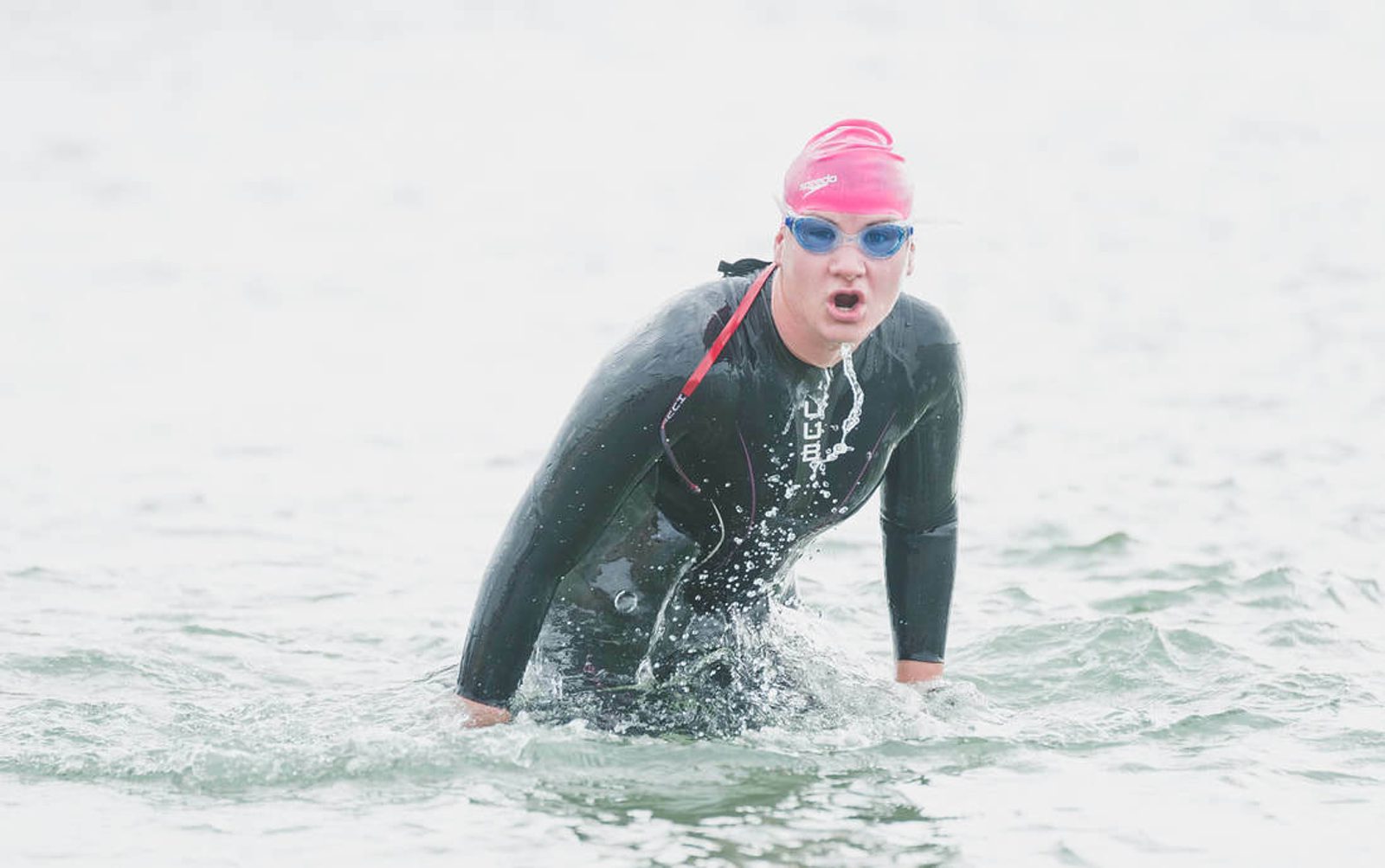 A swimmer wearing a black wetsuit, pink swim cap, and blue goggles emerges from the water. Water droplets are splashing around them as they push their way through the surface. The swimmer's mouth is open, possibly mid-breath or exhale.