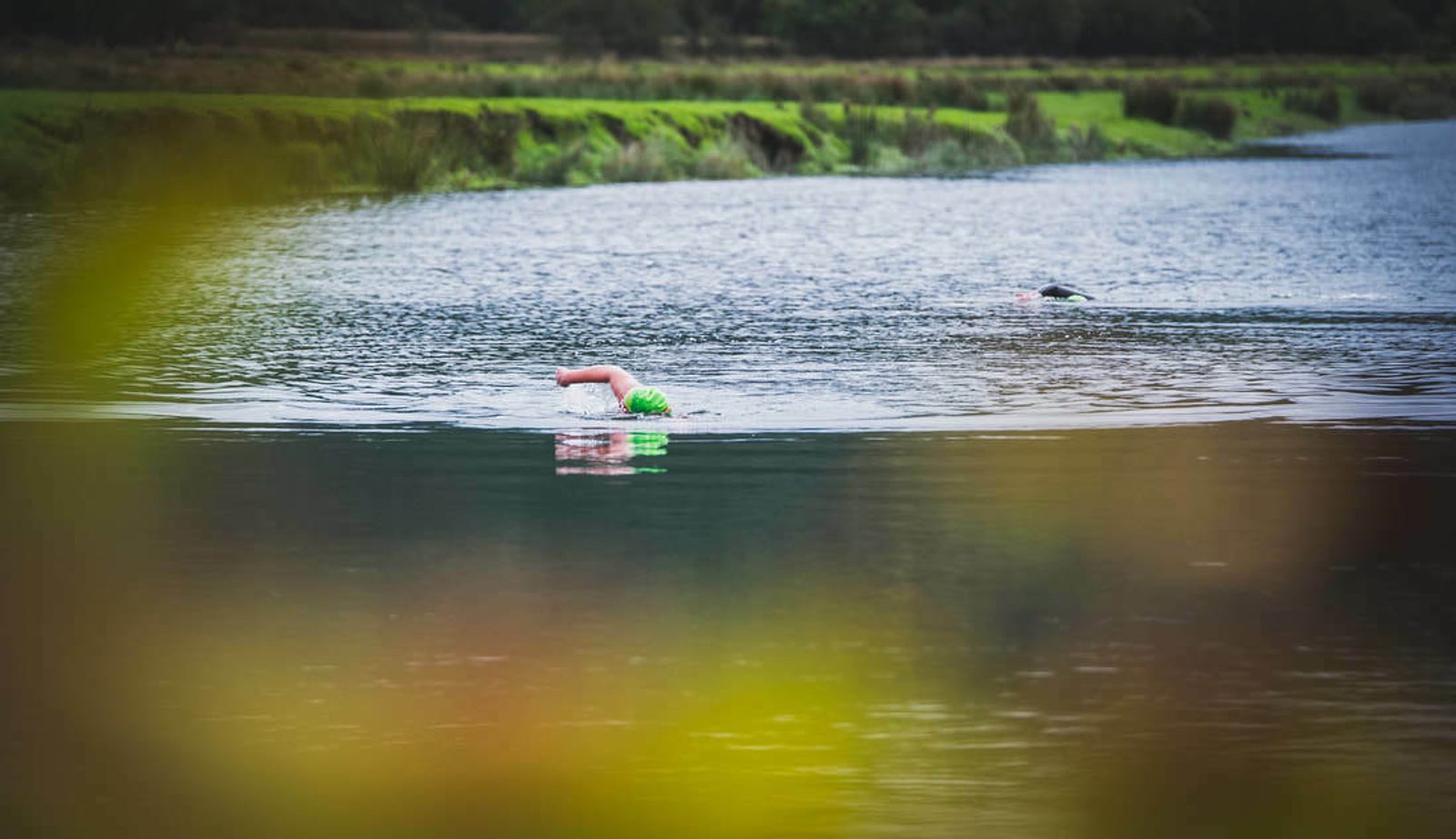 A person wearing a green swim cap and wetsuit is swimming in a body of water surrounded by lush greenery. Another swimmer is visible in the background. The image features blurred foliage in the foreground.