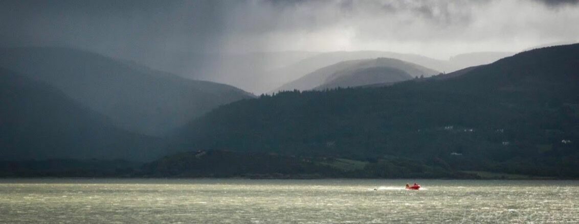 A red boat sails on a vast body of water under a cloudy, overcast sky. Dark hills and mountains loom in the background, partially shrouded in mist and rain, creating a dramatic and moody atmosphere.