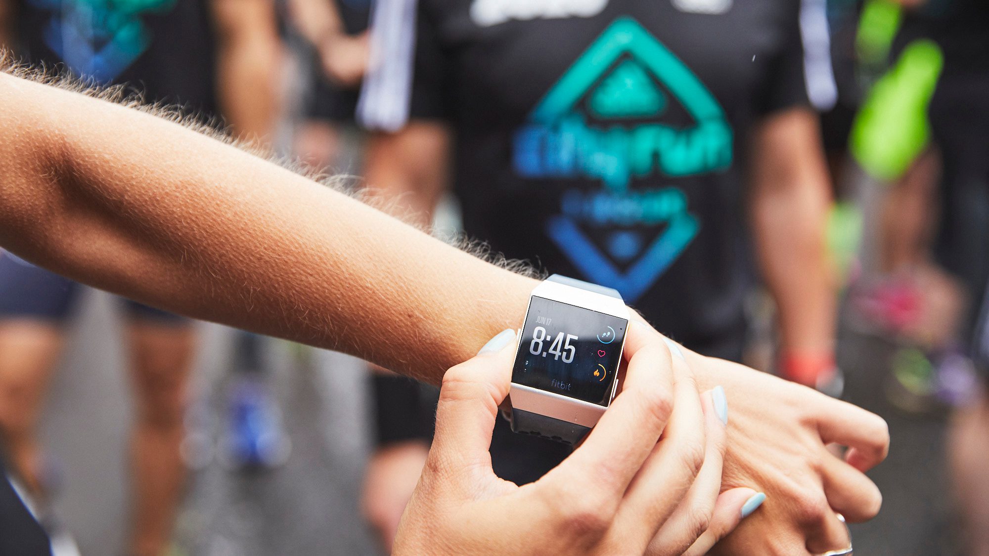 A runner checks the time on a smartwatch displaying 8:45. The background shows other runners in branded athletic wear, suggesting the scene is from a running event or race.