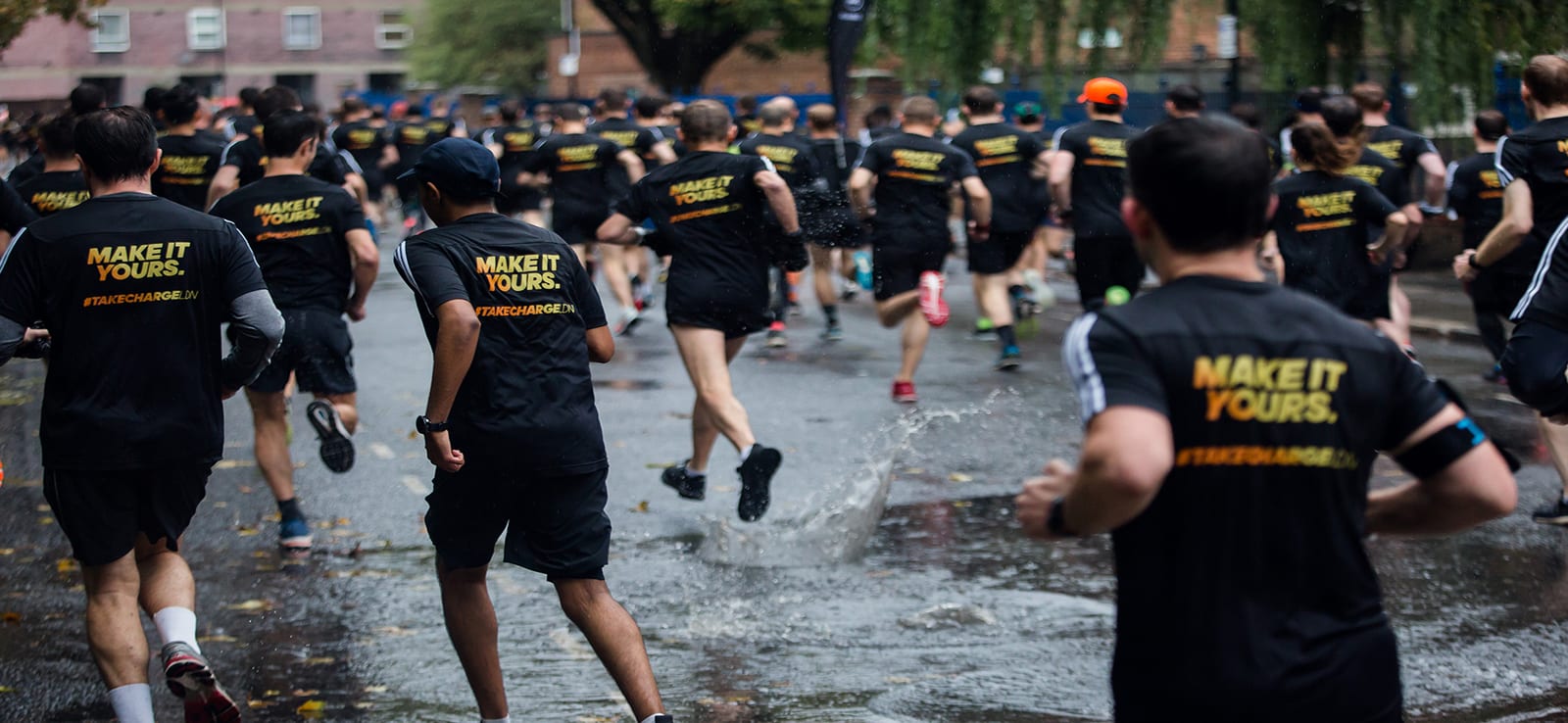 A group of runners, wearing black shirts with "MAKE IT YOURS" in yellow text on the back, participate in a race on a wet street. Several runners are mid-stride, splashing through puddles. Trees and buildings line the background.