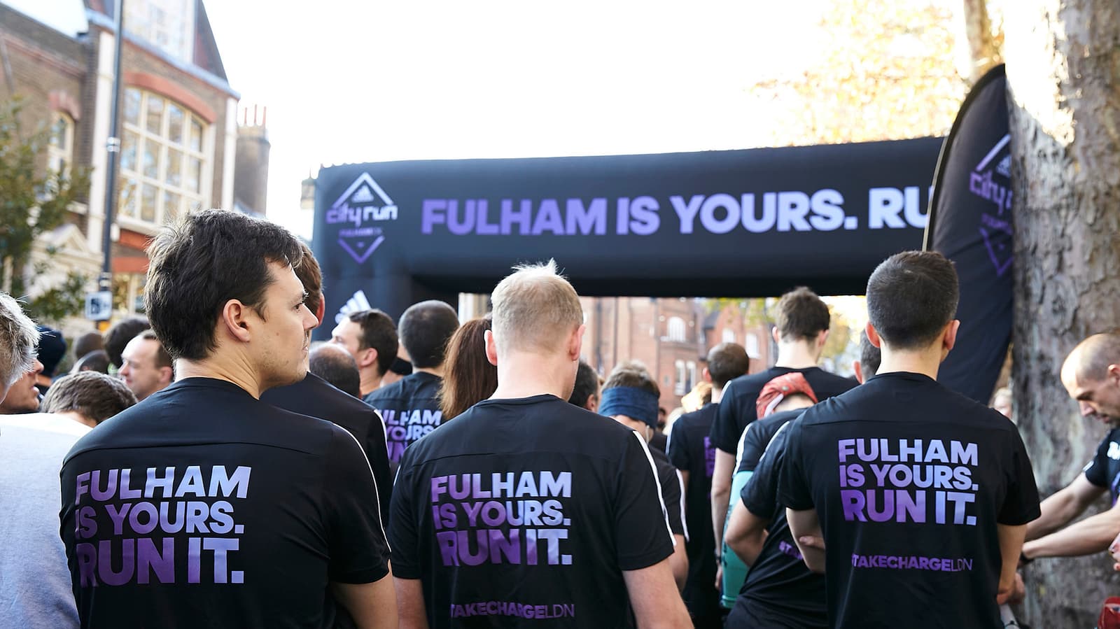 Participants wearing black shirts with the print "FULHAM IS YOURS. RUN IT." are gathered at the starting line of a running event. The event banner with the same slogan is visible in the background along with some trees and urban buildings.