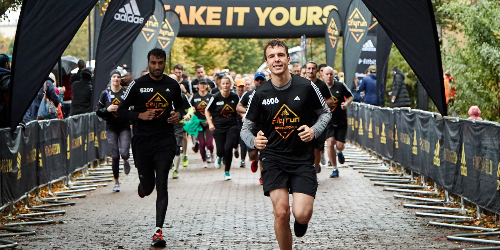 A group of runners participating in a race crosses the finish line. The runner in the foreground wears a black shirt with the number 4066. Surrounding him are other participants, and a large banner in the background reads “TAKE IT YOURS.” Bystanders line the side rails.