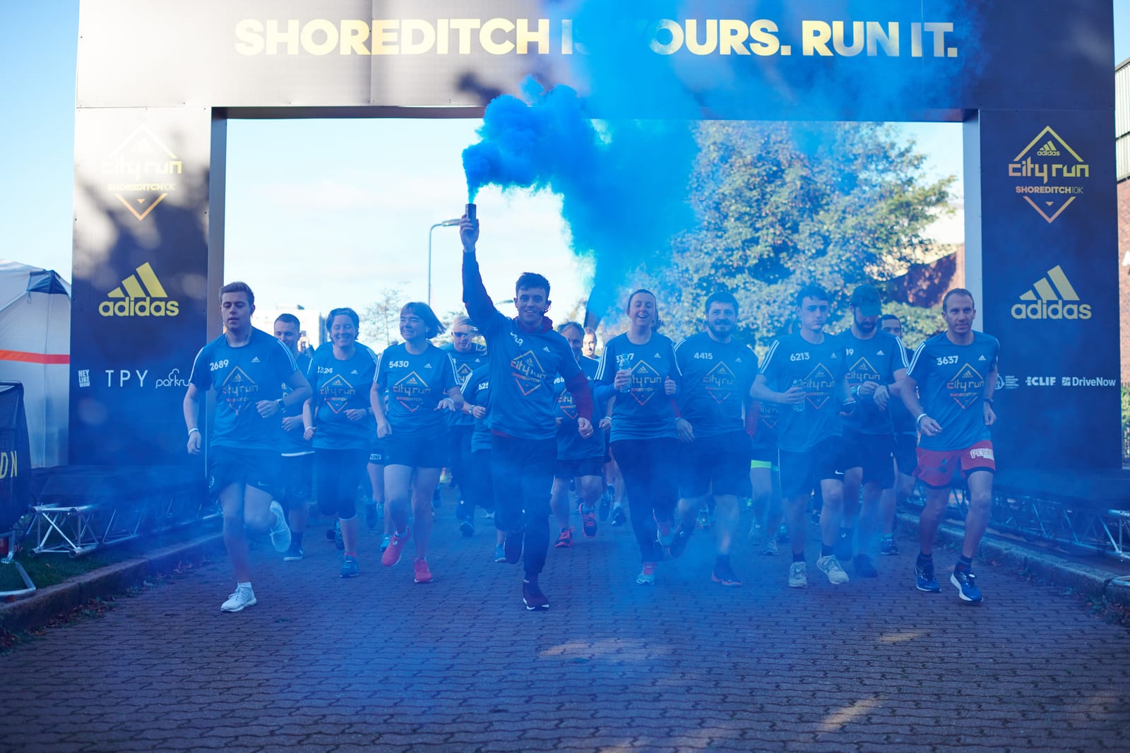 A group of enthusiastic runners, mostly dressed in blue athletic gear, start a race under an arch with the words "Shoreditch is Yours. Run It." One runner at the front holds a blue smoke flare, creating dramatic effects as they move forward.