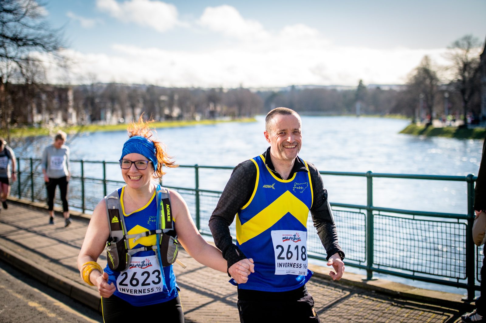 Two runners in blue and yellow athletic gear are competing in a race by a riverside. They are holding hands and smiling. The runner on the left wears glasses, a blue headband, and a race bib numbered 2623. The runner on the right wears a race bib numbered 2618.