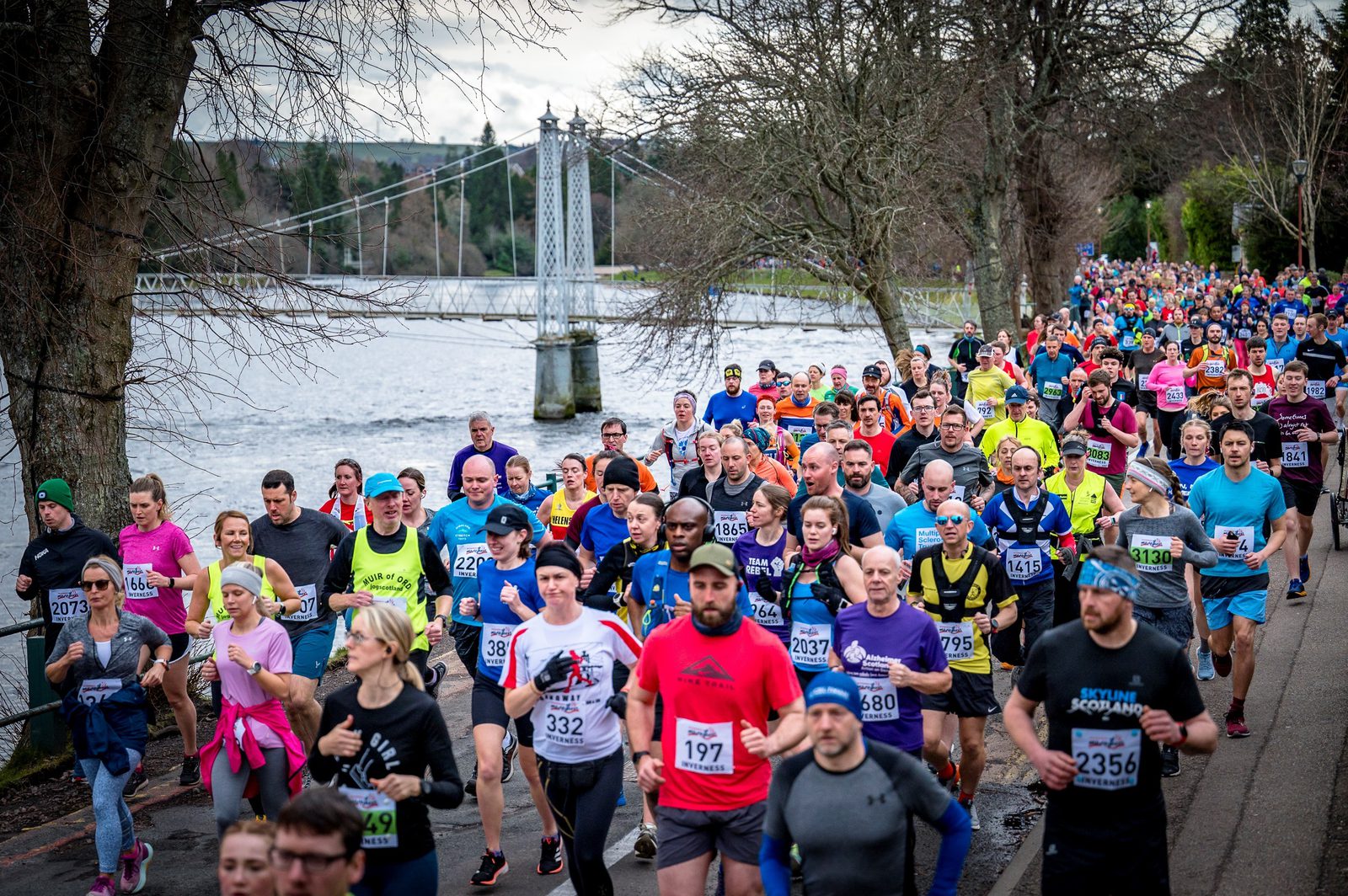 A large group of runners participating in a marathon or race alongside a river. The crowd includes people of various ages and genders wearing colorful athletic attire. In the background, there is a suspension bridge and a scenic, tree-lined path.