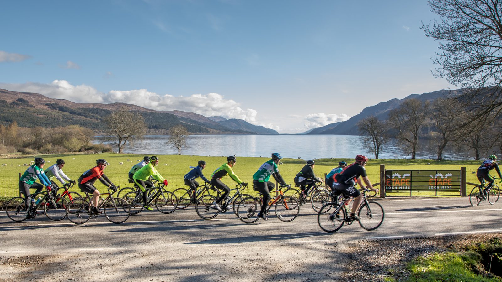 A group of cyclists in colorful gear riding on a paved road alongside a scenic lake. Mountains and clear sky are visible in the background. A banner reading "CAFΓ LOCH NES" can be seen to the right, with grassy areas and bare trees surrounding the lake.