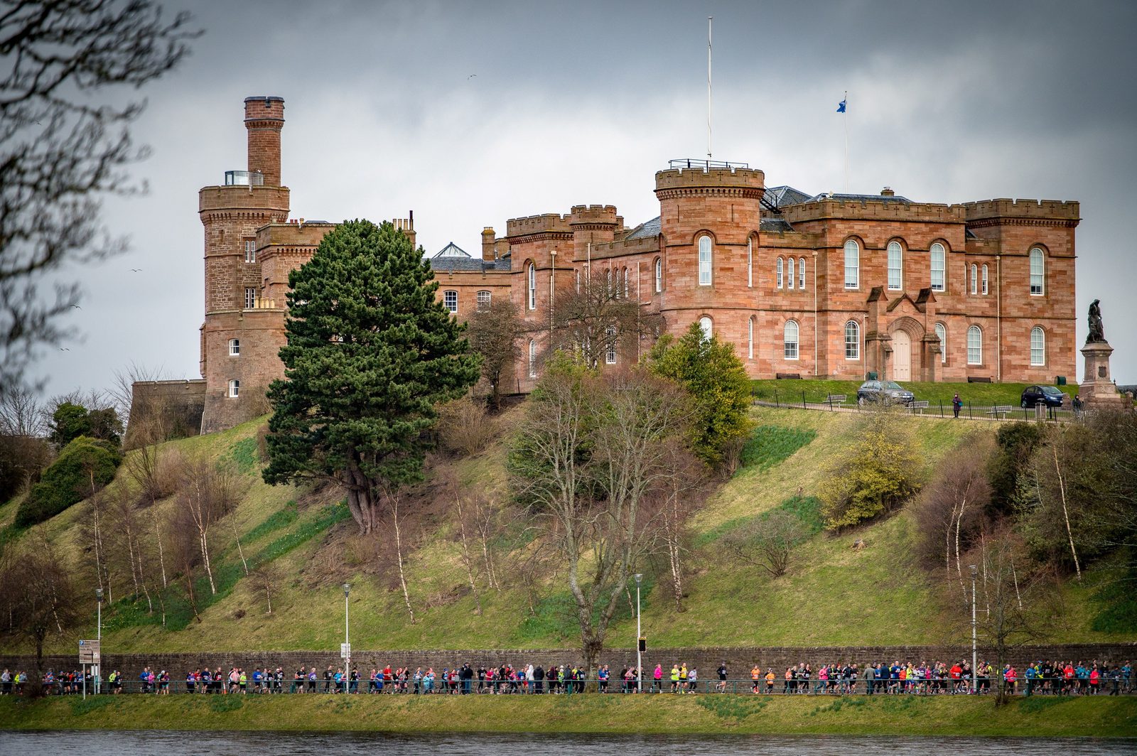 A large, historic castle made of reddish stone is situated on a grassy hill. Below the castle, a crowd of people lines a pathway near a body of water, possibly participating in an event or march. The sky is overcast, hinting at a cool, cloudy day.