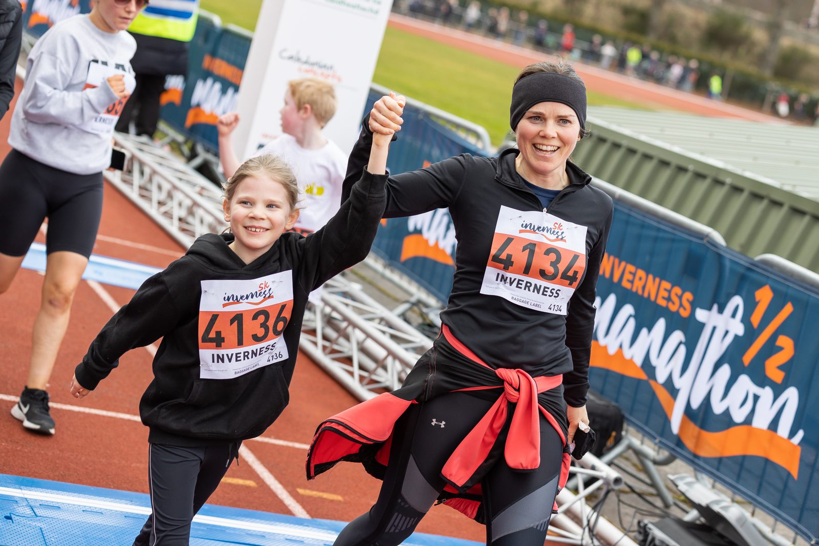 A woman and a child smiling and holding hands as they cross the finish line at a race. Both are wearing black running attire and race bibs, with the numbers 4134 and 4136. The background shows other participants and a banner that reads "Inverness 1/2 Marathon.