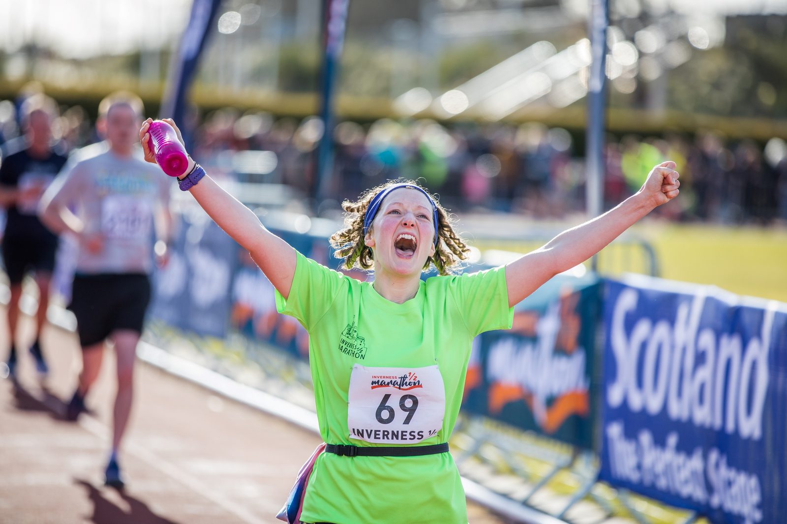 A marathon runner with number 69 on their shirt, wearing a green shirt and headband, raises their arms in celebration as they cross the finish line. They hold a pink bottle in one hand, while other runners can be seen in the background. The atmosphere is lively.