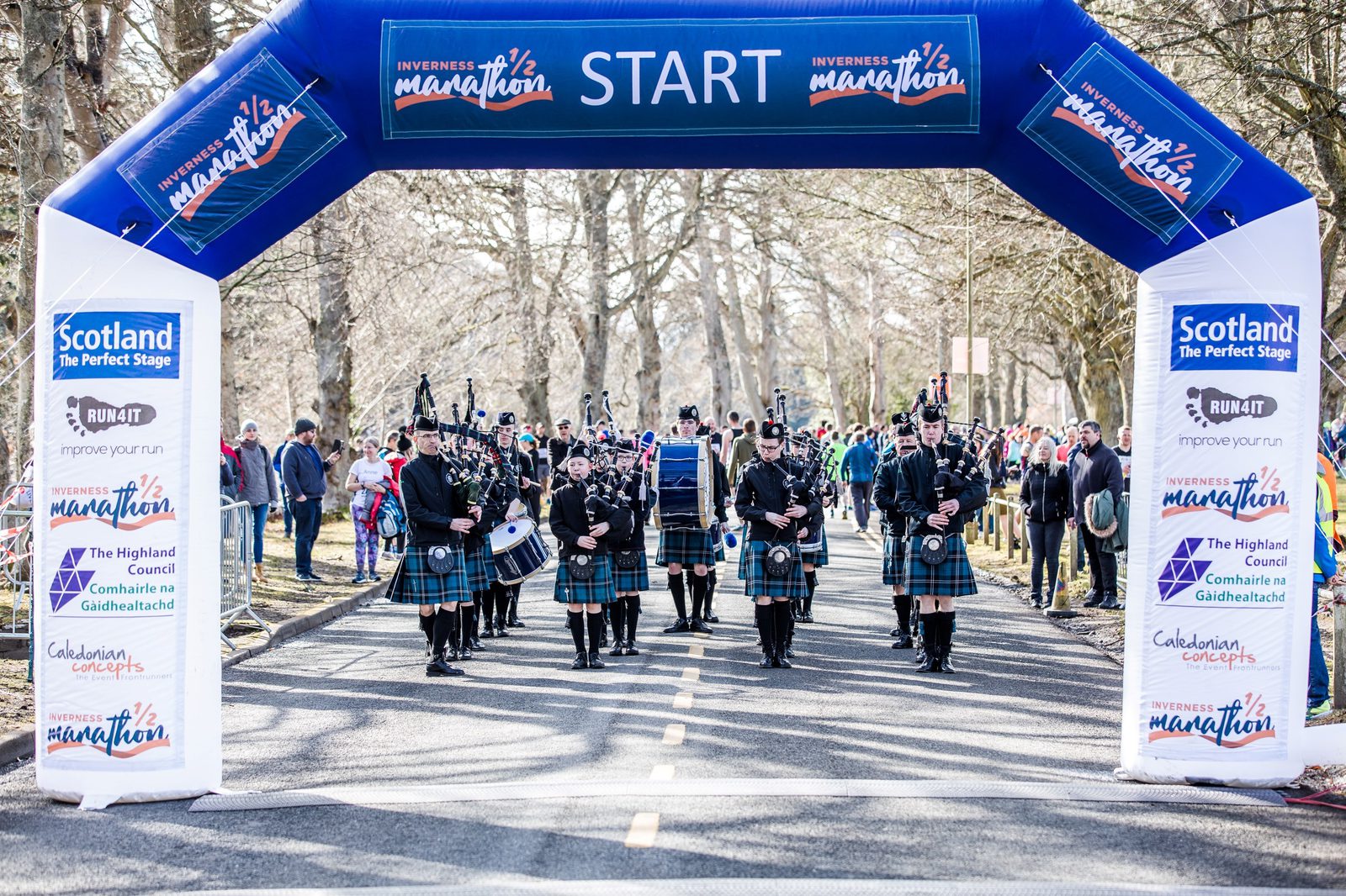 A group of kilted bagpipers lead the start of a marathon under a large blue inflatable arch that reads "START" and "Inverness marathon." Spectators line the road, surrounded by trees. Various sponsor logos are visible on the arch.