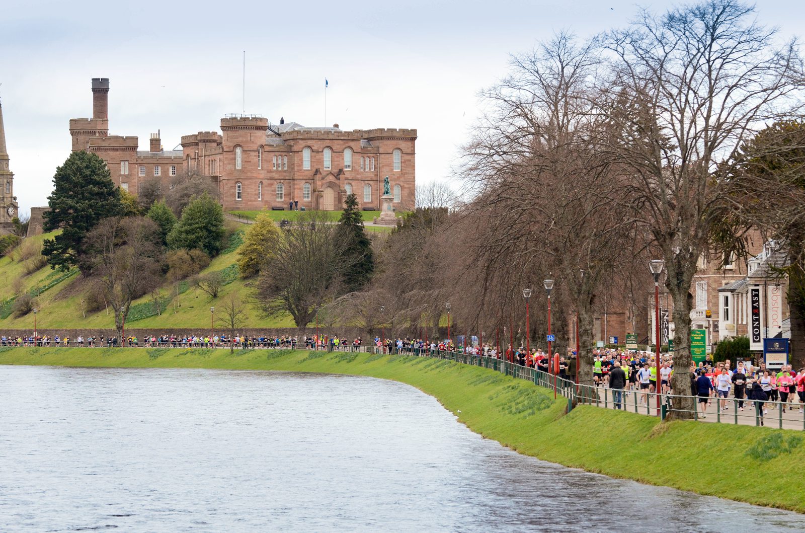 A large group of people are participating in a marathon along a riverside path. In the background, an expansive, historic castle sits atop a hill, surrounded by trees. The sky is overcast, and spectators line the path.