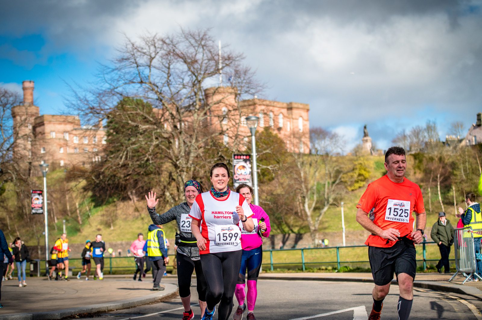 A group of runners participate in a race on a sunny day. They are running on a paved road with a park in the background, featuring leafless trees and a large building on a hill. Runners are wearing numbered race bibs, and some are smiling or waving at the camera.