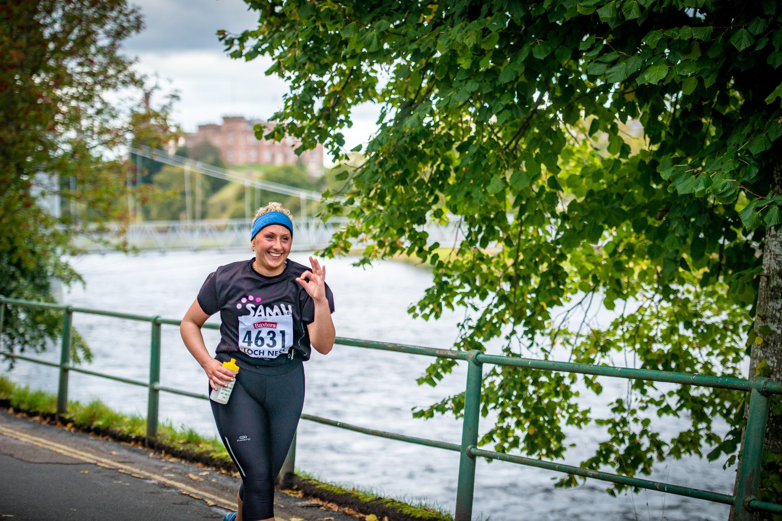 A woman runs along a riverside path, wearing a race number 4631. She is dressed in a black t-shirt, black leggings, and a blue headband. She holds a water bottle and smiles, giving a peace sign with her right hand. Lush green trees and a river are in the background.