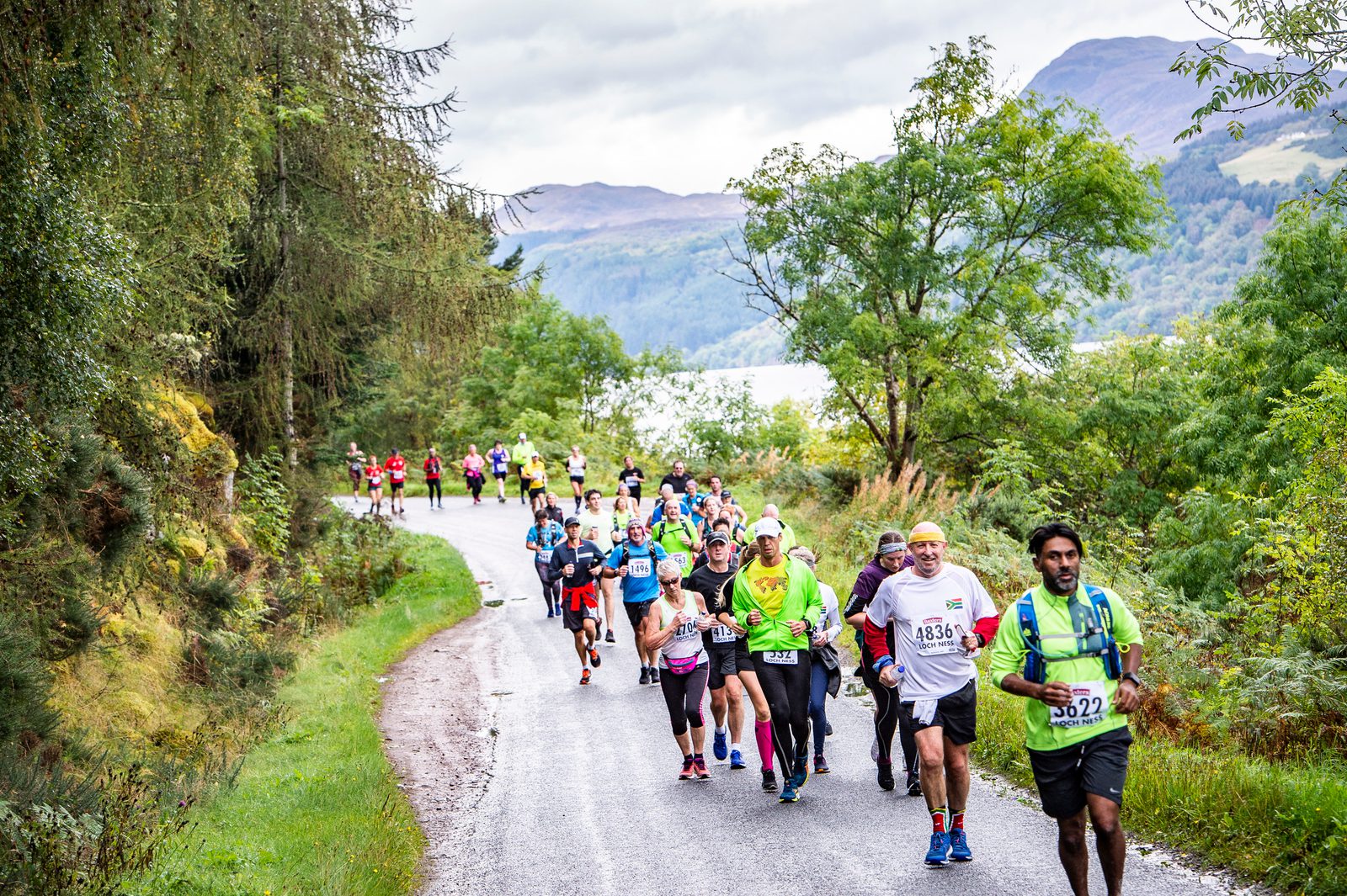 A group of runners partakes in a race on a scenic, tree-lined road. The participants, dressed in colorful athletic gear, include men and women of various ages. Lush greenery surrounds the road, with hills and a body of water visible in the background.