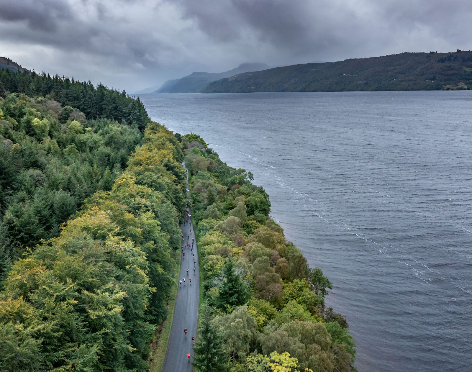 A scenic aerial view of a winding road flanked by lush green forests on one side and a large, tranquil body of water on the other. Small figures of cyclists can be seen riding along the road, set against a backdrop of misty mountains and a cloudy sky.