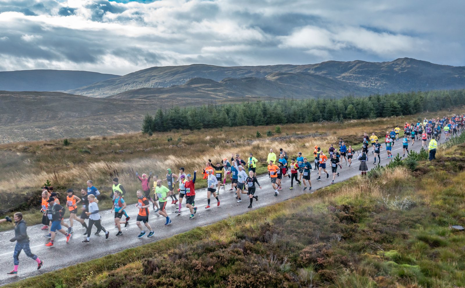 A large group of marathon runners in colorful athletic gear runs on a paved road through a scenic, mountainous landscape with grassy plains, meadows, and trees. The sky is overcast with patches of blue, and the road stretches into the distance.