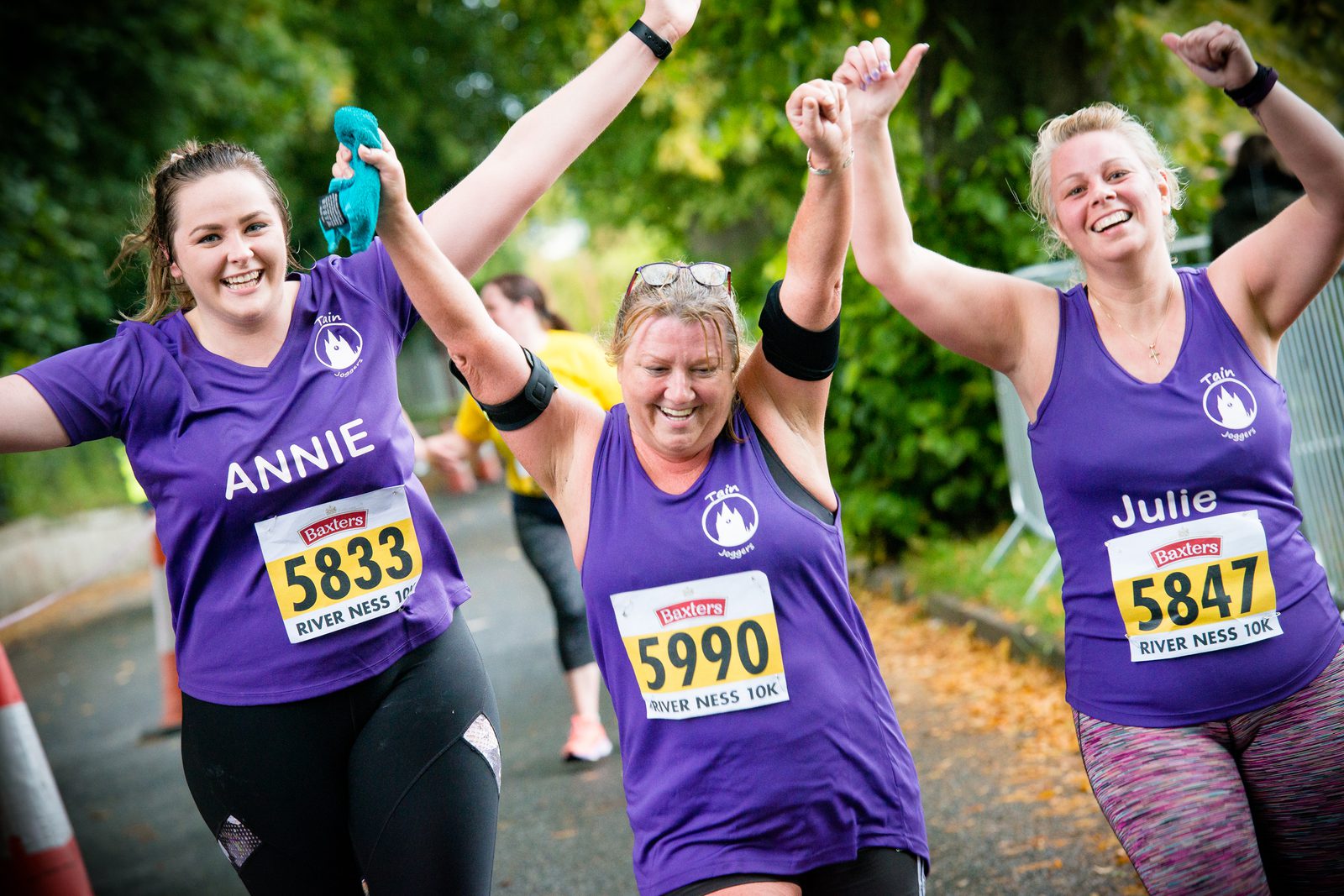 Three women in purple shirts celebrate as they near the finish line of a race, wearing bib numbers 5833, 5990, and 5847. They joyfully raise their arms in triumph. The race bib for one participant reads "River Ness 10K," and the names on the shirts are visible.