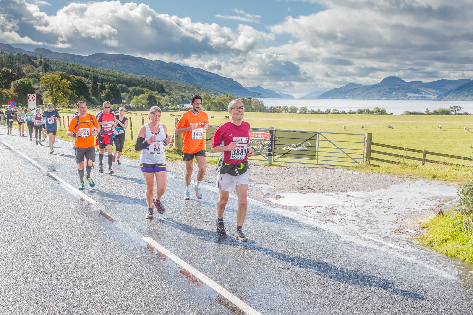 A group of runners is participating in an outdoor marathon on a scenic road beside a lake. The sky is partly cloudy, and there are hills and greenery in the background. Bib numbers are visible, with one runner in a maroon shirt leading the group.