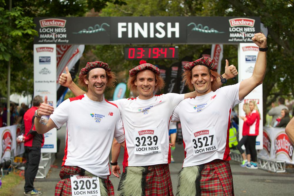 Three men wearing white shirts, kilts, and red Scottish hats pose with thumbs up after finishing the Baxters Loch Ness Marathon. They stand in front of a large finish line banner with a clock displaying their time of 3 hours, 19 minutes, and 47 seconds.