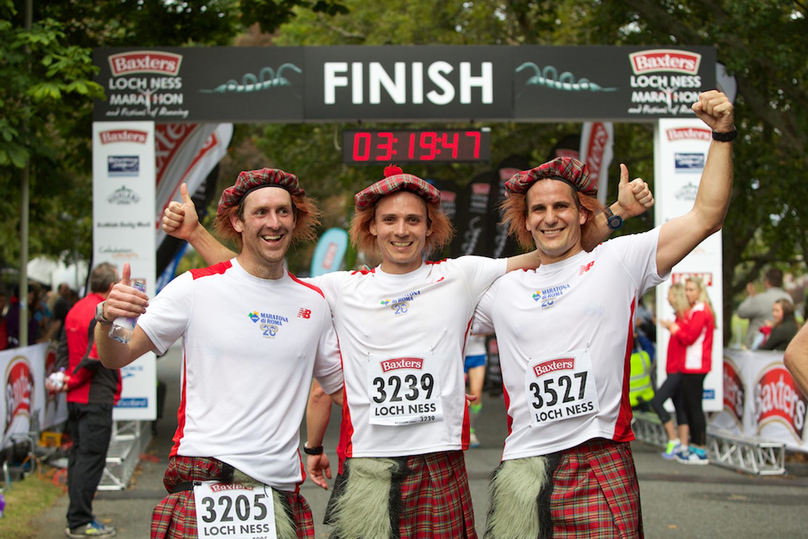 Three men wearing white shirts, kilts, and red Scottish hats pose with thumbs up after finishing the Baxters Loch Ness Marathon. They stand in front of a large finish line banner with a clock displaying their time of 3 hours, 19 minutes, and 47 seconds.