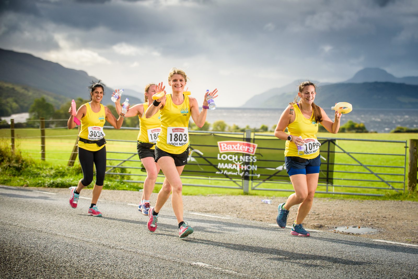 Four women running in a road race, all wearing yellow running shirts. They are smiling and waving, with race numbers pinned to their shirts. The background features a scenic view of mountains and a lake under a cloudy sky. A banner reads "Baxters Loch Ness Marathon.